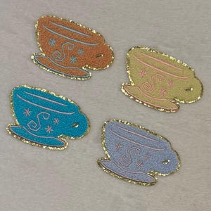 May include: Four embroidered teacup patches in shades of orange, yellow, blue, and light blue. Each patch has a gold glitter border and features a decorative design with pink and blue accents. The patches are on a white, textured surface.