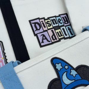 May include: A white canvas tote bag featuring embroidered patches including white cartoon gloves forming a heart shape, a rectangular patch with the text Disney Adult in pastel colors, and a blue wizard hat with stars and a crescent moon.