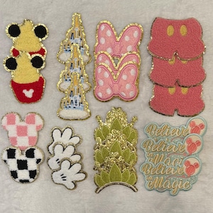 May include: A collection of colorful, embroidered patches featuring iconic Disney designs. Includes Mickey Mouse ears, castle, Minnie Mouse bows, shorts, hands, and text that says "Believe in Magic". Each patch has a gold glitter border.