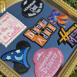 May include: A collection of embroidered patches displayed on a mirror with a gold frame. Patches include a black oval with white text, a pink stamp-shaped patch with a castle, a blue wizard hat, a glittery pink heart, and orange and black text patches.