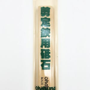 May include: A rectangular sharpening stone in a clear plastic sleeve. The stone is light brown, and the sleeve has green Japanese text and the brand name "Oka-tuna" printed on it. The packaging is sealed with a metal rivet.