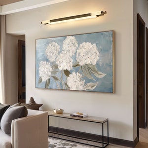 May include: A light blue canvas painting with white hydrangea flowers. The painting is framed in a gold frame and is hanging on a white wall above a black metal console table.