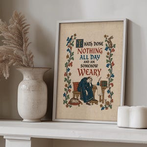 Op de afbeelding: Een ingelijste kunstdruk met een middeleeuwse illustratie van een persoon in een blauwe mantel die aan een bureau met een kat zit, omringd door bloemenranden en de tekst: T HATH DONE NOTHING ALL DAY AND AM SOMEHOW WEARY in rood en blauw.