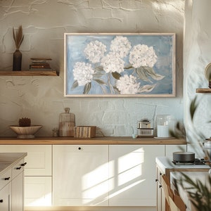 May include: A framed painting of white hydrangeas on a blue background. The painting is hanging on a white wall in a kitchen.
