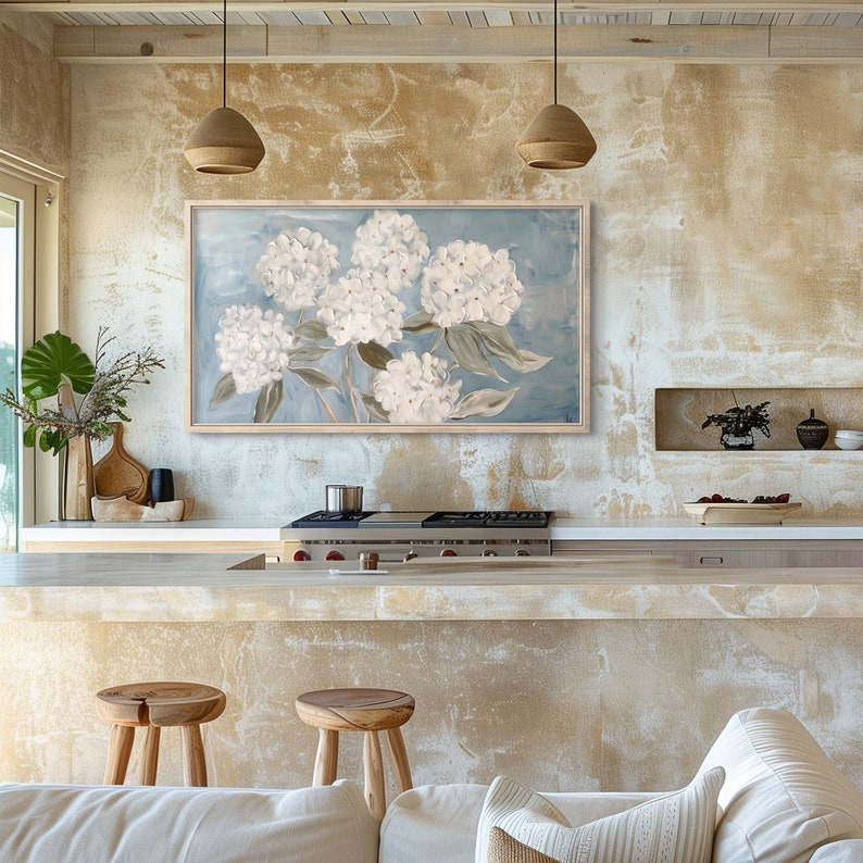 May include: A framed painting of white hydrangeas on a blue background, hanging on a wall with a textured, beige finish. The painting is above a kitchen counter with a stainless steel stovetop and a white countertop.