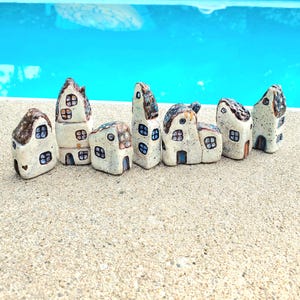 Puede incluir: Una colección de siete casas en miniatura de cerámica con diferentes formas y tamaños. Cada casa presenta una base de color crema con detalles marrones y azules en el tejado y alrededor de las ventanas y puertas. Las casas están dispuestas sobre una superficie clara con un fondo azul.
