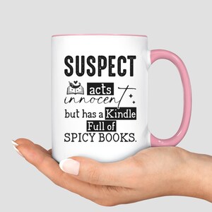 May include: White ceramic mug with a pink rim and handle. The mug has black text that reads "SUSPECT acts innocent but has a Kindle Full of SPICY BOOKS."