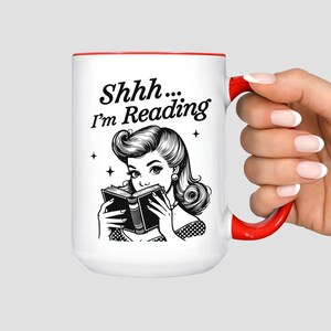 May include: White ceramic mug with a red interior and handle. The mug features a black and white illustration of a woman reading a book, with the text "Shhh... I'm Reading" in black script. A perfect gift for book lovers.
