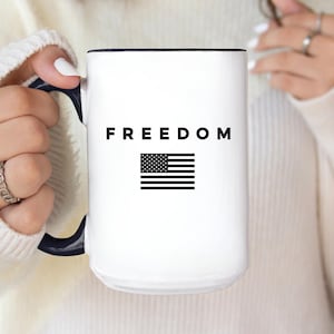 May include: White ceramic mug with a black handle and rim. The mug features the word "FREEDOM" in black block letters above a black and white American flag design. A simple and patriotic design.