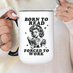 May include: White ceramic mug with a black interior and handle. The mug features the text "BORN TO READ FORCED TO WORK" with a black and white illustration of a woman reading a book and holding a cup. The mug is held by a person.