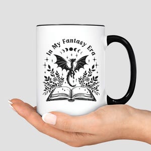 May include: White ceramic mug with a black handle and rim. The mug features a black and white illustration of a dragon, open book, and the text "In My Fantasy Era". The design is surrounded by floral and celestial elements.