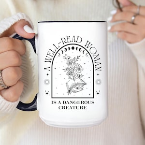 May include: White ceramic mug with a black interior and handle. The mug features the text "A WELL-READ WOMAN IS A DANGEROUS CREATURE" in black, along with a floral and book design. The design is centered on the mug.