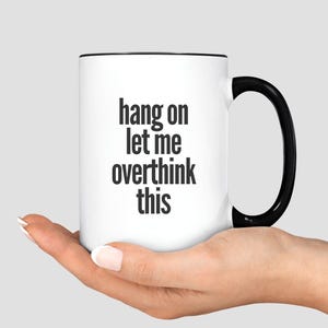 May include: White ceramic mug with a black interior and handle. The mug features the text "hang on let me overthink this" in black, centered on the front. The mug is held in a person's hand.