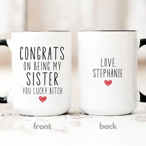 May include: Two white ceramic mugs with black handles and rims. One mug reads "CONGRATS ON BEING MY SISTER YOU LUCKY BITCH" with a red heart. The other mug says "LOVE, Stephanie" with a red heart. The mugs are on a light wood surface.