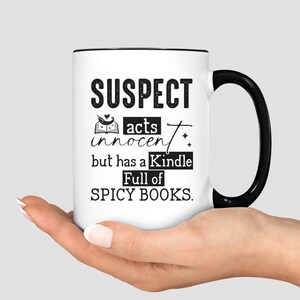 May include: White coffee mug with a black rim and handle. The mug has black text that reads "SUSPECT acts innocent but has a Kindle Full of SPICY BOOKS."