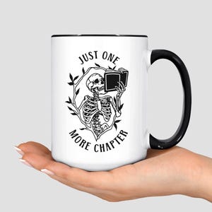 May include: White ceramic mug with a black interior and handle. The mug features a black and white illustration of a skeleton reading a book, surrounded by a leafy design. Text on the mug reads "JUST ONE MORE CHAPTER."