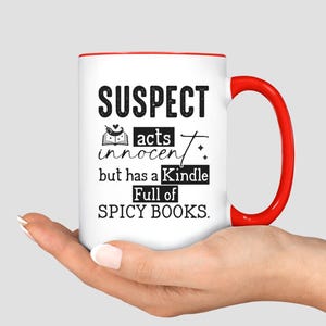 May include: White coffee mug with a red rim and handle. The mug has black text that reads "SUSPECT acts innocent but has a Kindle Full of SPICY BOOKS."