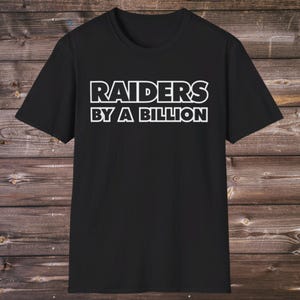 Puede incluir: Camiseta negra con la frase blanca "RAIDERS BY A BILLION". El texto está en negrita y delineado. La camiseta tiene cuello redondo y mangas cortas. El fondo es una superficie de madera.
