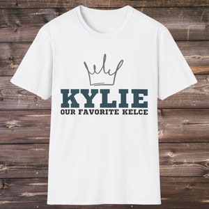 Puede incluir: Camiseta blanca con un dise&ntilde;o de corona sobre el nombre "KYLIE" en letras de bloque verde azulado, con "OUR FAVORITE KELCE" debajo. La camiseta se muestra sobre un fondo de madera.