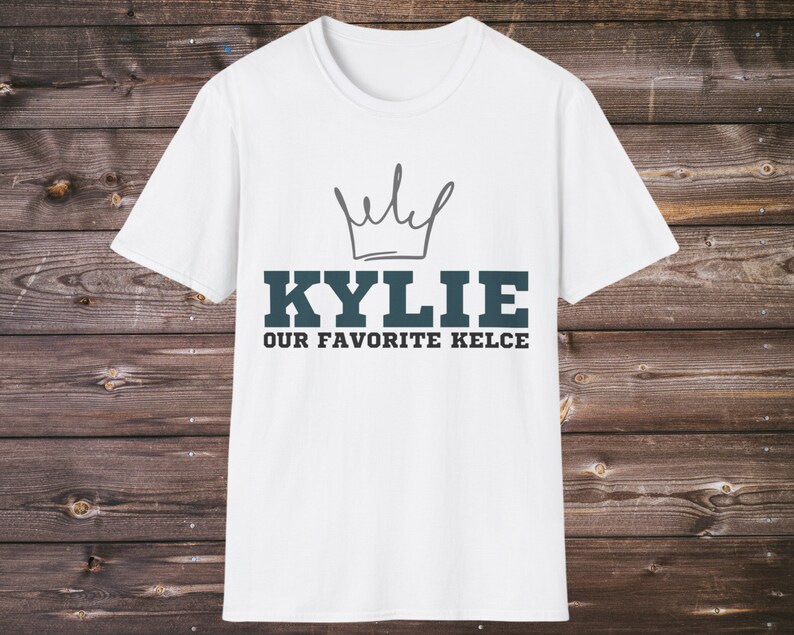 Puede incluir: Camiseta blanca con un dise&ntilde;o de corona sobre el nombre "KYLIE" en letras de bloque verde azulado, con "OUR FAVORITE KELCE" debajo. La camiseta se muestra sobre un fondo de madera.