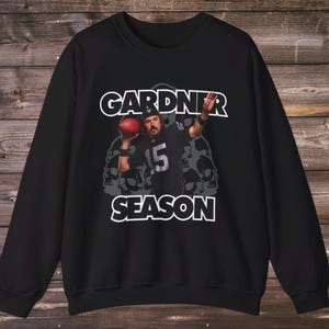 Puede incluir: Sudadera negra con un gráfico de un jugador de fútbol americano sosteniendo un balón. El texto "GARDNER SEASON" está impreso en blanco encima y debajo de la imagen. El jugador lleva una camiseta negra con el número 15.