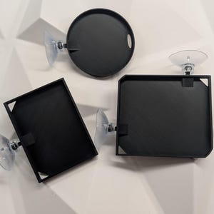 May include: Three black trays with textured surfaces and suction cups. One tray is round with a handle, and the other two are rectangular with square corners. The trays are designed to attach to a smooth surface.