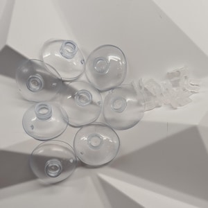 May include: A selection of clear, circular suction cups with central holes and a few small, clear plastic clips. The suction cups are arranged in a white, geometric container.