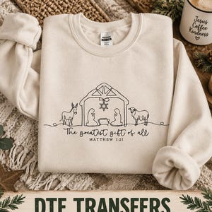 May include: A sand-colored crewneck sweatshirt featuring a black line-art graphic of a nativity scene with a donkey, a sheep, and the text "The greatest gift of all" followed by "MATTHEW 1:21" printed in a simple, clean, dark-colored font.