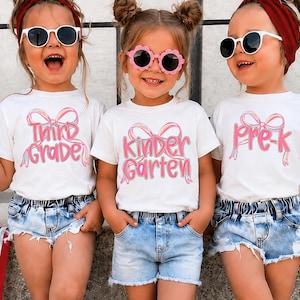 May include: Three white t-shirts featuring pink and blue bow graphics with the text Third Grade, Kindergarten, and Pre-K. The shirts are paired with light blue denim shorts, white high-top sneakers, and socks with a red, white, and blue star and stripe pattern.