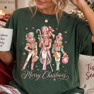 May include: A dark green short-sleeve t-shirt featuring a graphic of three nutcracker figures in pink and teal uniforms with gold accents. The text "Merry Christmas" is printed below the figures in a cursive font. The shirt is made of cotton.