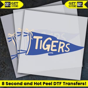 May include: A blue pennant-shaped DTF transfer featuring the word TIGERS in large cream-colored letters. The design includes a cream-colored ribbon tied at the left side. The image includes text stating 55% GET 3 ITEMS, 60% GET 10 ITEMS, and 8 Second and Hot Peel DTF Transfers.
