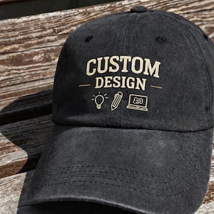 May include: A black baseball cap with the words "CUSTOM DESIGN" in a tan color. Below the text are three small icons: a lightbulb, a pencil, and a laptop. The cap is made of a soft material and has a curved brim.