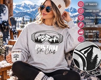 Puede incluir: Una sudadera gris claro de cuello redondo con un gráfico negro de gafas de esquí que contienen un paisaje de montaña y bosque, con el texto CUSTOM Ski Club impreso debajo. El diseño se muestra como una transferencia DTF sobre tela.