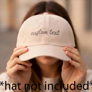 May include: A beige baseball cap featuring the words custom text embroidered in a dark gray cursive font across the front panels. The cap has a structured crown, a curved brim, and a button at the top center, showcasing a classic six-panel design.