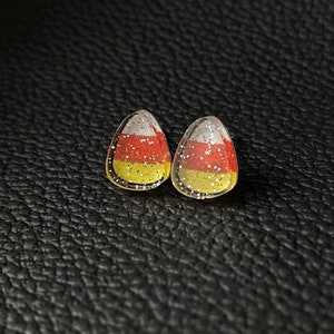May include: A pair of candy corn-shaped stud earrings. Each earring features a yellow base, a red middle section, and a white top with silver glitter. The earrings are set against a dark, textured background.