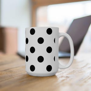 May include: A white ceramic mug featuring a repeating pattern of solid black polka dots across the exterior surface. The mug has a cylindrical body and a curved handle on the right side, designed for holding hot or cold beverages in a simple style.