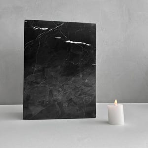 May include: A black marble slab with white veining sits on a white surface next to a white candle with a lit flame.