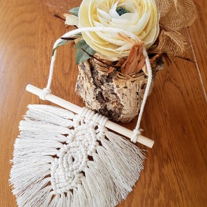 Könnte beinhalten: Ein weißer Makramee-Wandbehang mit einem Holzstab und Fransen. Der Behang hat die Form eines Blattes und ein kreisförmiges Muster in der Mitte.