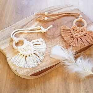 Può includere: Due arazzi da parete in macramè con anelli di legno e frange. Gli arazzi sono su una superficie di legno. Uno è bianco e l'altro è marrone.