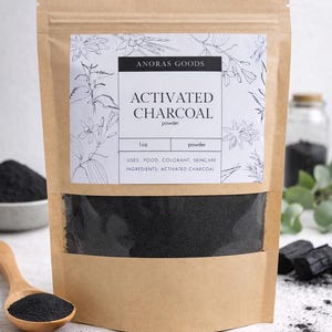 May include: A brown paper bag with a clear panel showing black activated charcoal powder. The label says "ANORAS GOODS ACTIVATED CHARCOAL powder" with botanical drawings. A wooden spoon holds a portion of the black powder.