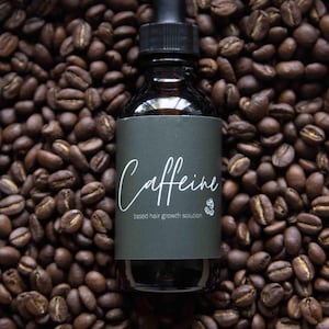 May include: A dark glass bottle with a dropper, labelled "Caffeine based hair growth solution." The bottle rests on a bed of roasted coffee beans. The label is dark green with white script.