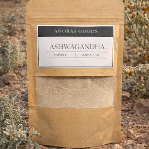 Puede incluir: Una bolsa de papel marr&oacute;n con polvo de ashwagandha. La bolsa tiene una ventana transparente que muestra el polvo marr&oacute;n claro. La etiqueta dice "ANORAS GOODS" y "ASHWAGANDHA" con "POWDER" y "SAMPLE &frac12; OZ" debajo.