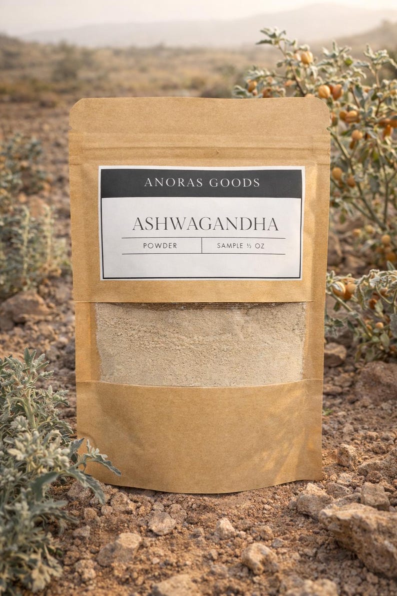 Puede incluir: Una bolsa de papel marr&oacute;n con polvo de ashwagandha. La bolsa tiene una ventana transparente que muestra el polvo marr&oacute;n claro. La etiqueta dice "ANORAS GOODS" y "ASHWAGANDHA" con "POWDER" y "SAMPLE &frac12; OZ" debajo.