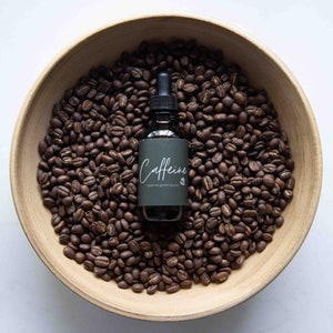 Puede incluir: Una botella de vidrio oscuro con un gotero, llena de un líquido oscuro. La botella está etiquetada como "Caffeine Scalp Hair Growth Solution" y está sentada sobre un lecho de granos de café en un tazón de madera.