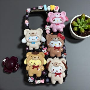 May include: A black phone case adorned with cartoon character figures dressed as bears. The word "LOVE" is spelt out in colourful letters at the top. The case also features pearls and small sweet decorations.