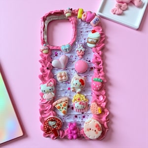 K&ouml;nnte beinhalten: Eine rosa Handyh&uuml;lle, verziert mit verschiedenen 3D-Verzierungen. Die H&uuml;lle hat einen ger&uuml;schten Rand und ist mit Miniatur-Cupcakes, Herzen, Hello Kitty-Figuren, einem Pizzast&uuml;ck und einer Kaffeetasse verziert. Der Hintergrund ist hellrosa.