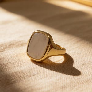 May include: A gold-colored ring with a rectangular, light-colored stone. The ring is set on a beige surface, with a gold-colored mirror and other objects in the background. The ring's design is simple and elegant.