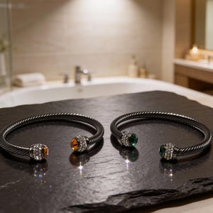May include: Two silver-tone bracelets with a twisted design. Each bracelet features a decorative end with a central gemstone, one orange and one green, surrounded by clear crystals. The bracelets are displayed on a dark, textured surface.