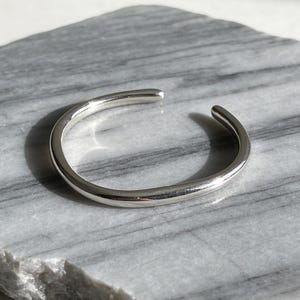 May include: A silver-colored, open-ended cuff bracelet. The bracelet is smooth and rounded, with a simple, minimalist design. It is resting on a gray and white marble surface, with natural light casting shadows.