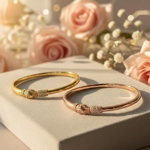 May include: Two elegant bangles, one gold-toned and one rose gold-toned, rest on a neutral surface. Each bracelet features a decorative knot design embellished with small, sparkling stones. Soft pink roses and delicate white flowers provide a romantic backdrop.